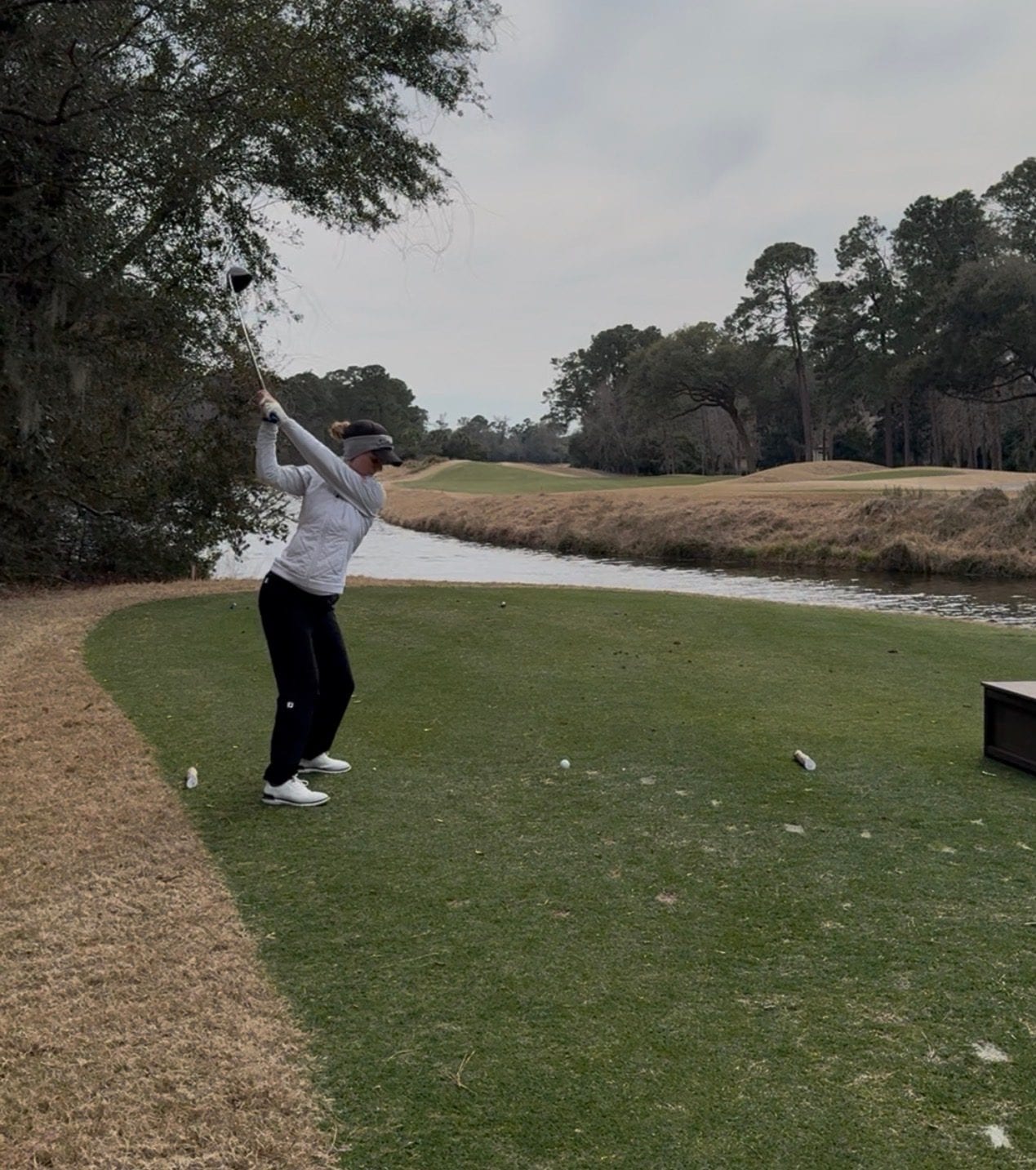 Sofie mid-swing with a driver, hitting a tee shot over water.